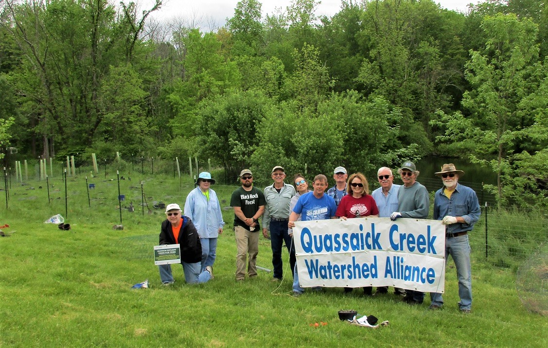 A group of volunteers hold a sign "Quassaick Creek Watershed Alliance" while standing in front of an area recently planted with small trees