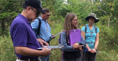 Four community volunteers stand in a grassy area holding clipboards during an outdoor training. By L Heady