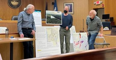 Three men looking at maps and a large photo of a waterbody at a Town of Mount Pleasant NRI committee meeting.