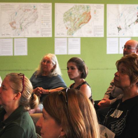 Audience of community members at NRI meeting with maps hanging in the background