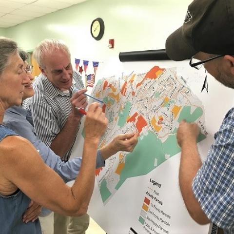 Three people gathered around map on easel, pointing out and discussing features on the map. By L Heady