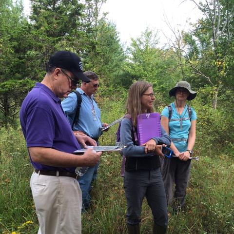 Four community volunteers stand in a grassy area holding clipboards during an outdoor training. By L Heady