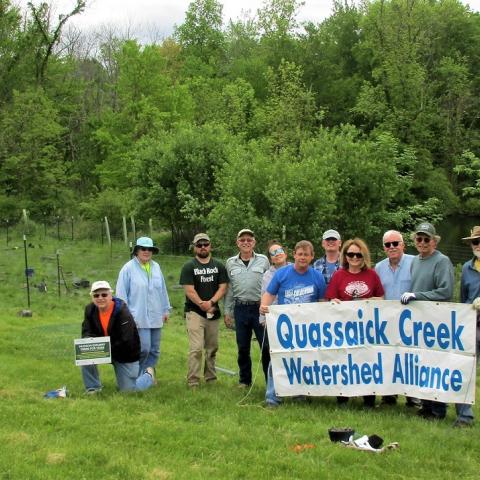 A group of volunteers hold a sign "Quassaick Creek Watershed Alliance" while standing in front of an area recently planted with small trees