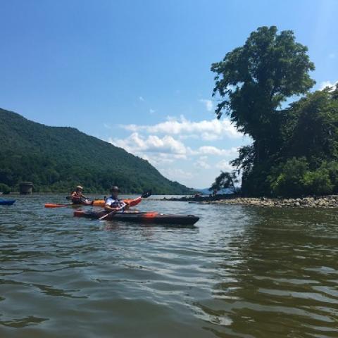 Kayakers paddle on the Hudson River through the Hudson Highlands, under a blue sky.